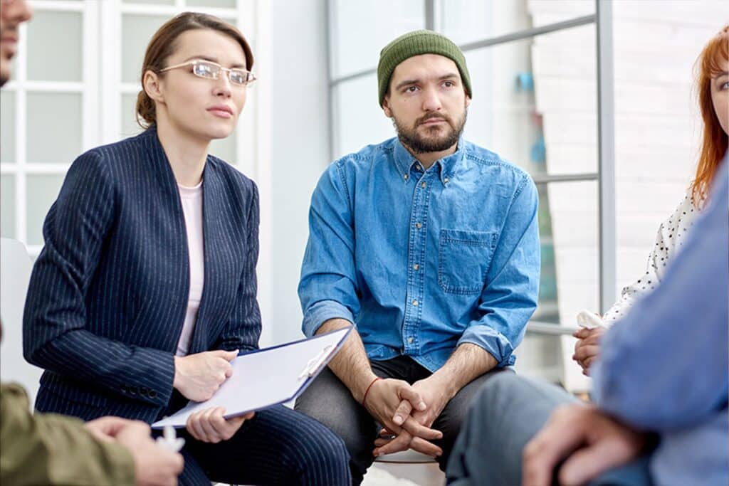 Halfway house group featuring two women wearing blouses, man wearing denim shirt and beanie, and a therapist with glasses and pinstripe suit